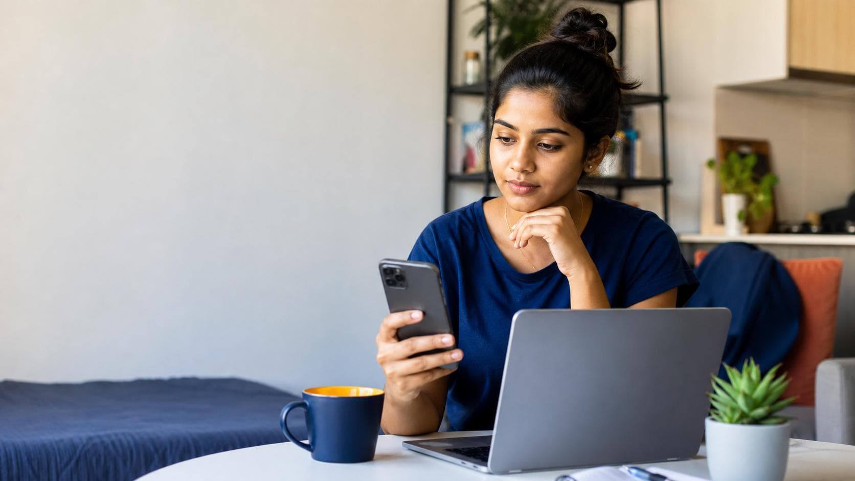 Sri Lankan-Australian student comparing mobile plan options on a phone beside a laptop in a realistic apartment